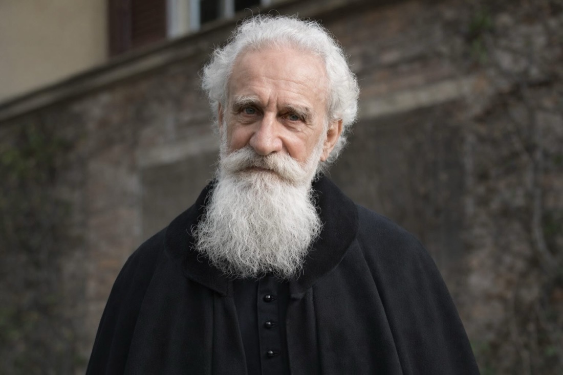 Mgr Ghika en manteau noir, devant un mur de pierre.